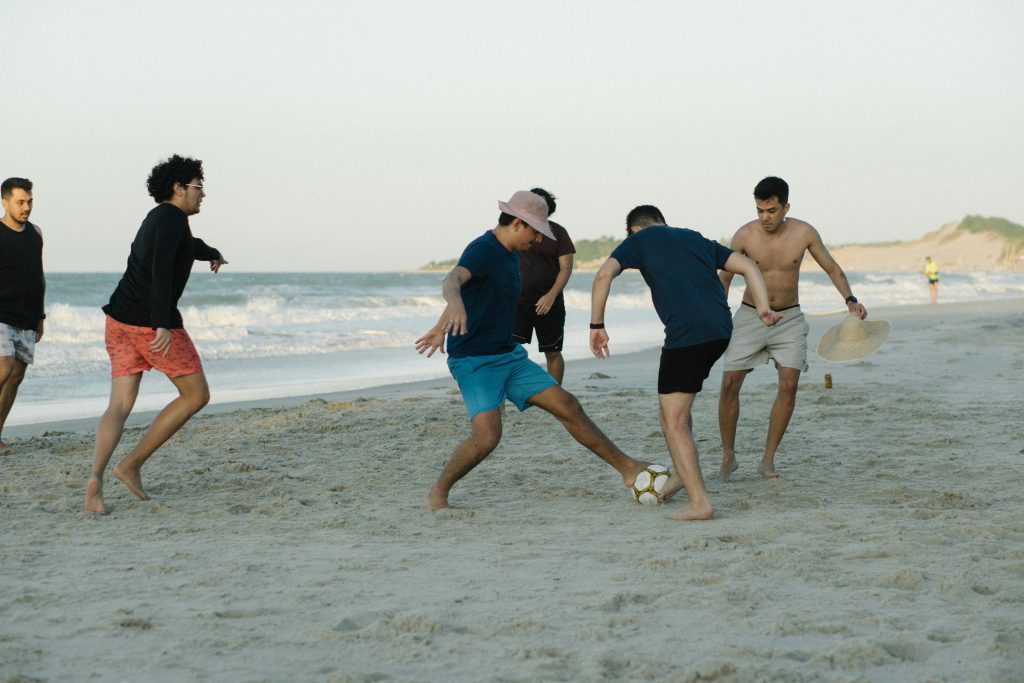 Five young adults playing soccer on a sunny beach, enjoying leisure and sports.