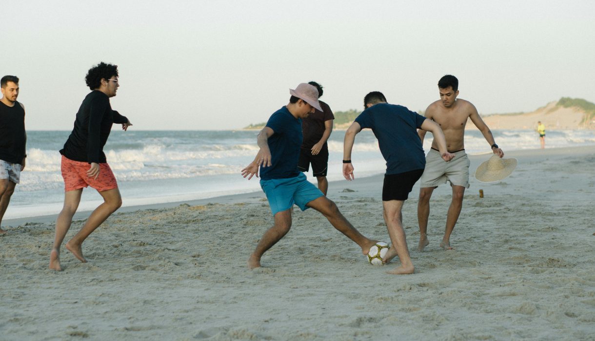 Five young adults playing soccer on a sunny beach, enjoying leisure and sports.