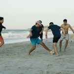 Five young adults playing soccer on a sunny beach, enjoying leisure and sports.